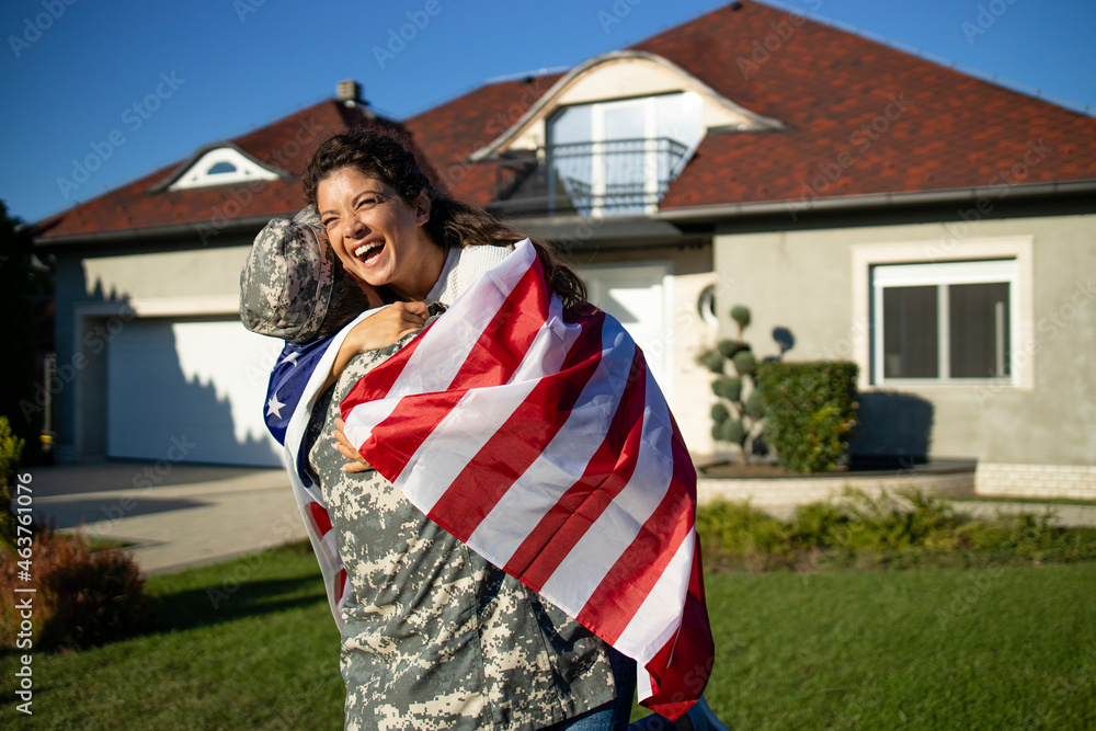 Soldier in uniform coming home and his lovely wife with American flag running into his arms celebrating reunion.